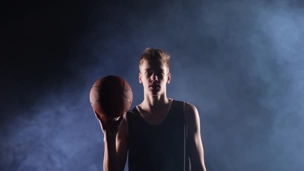 Um jogador do sexo masculino gira um basquete em seu dedo em câmera lenta. Jovem jogador de basquete posando em um estúdio fumegante escuro sob os holofotes. Fechar. — Vídeo de Stock