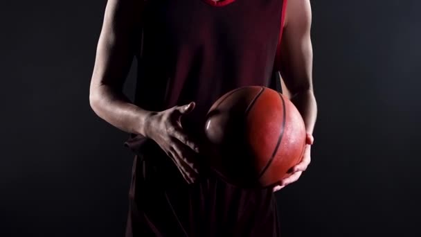 Ung basketspelare i en mörk studio på en svart bakgrund. Närbild av en idrottare hand, hålla en basket, rulla den, kasta den från en hand till en annan. Långsamma rörelser. — Stockvideo