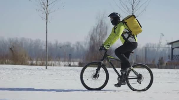 Ekspresowa dostawa żywności na rowerze. Mężczyzna z żółtym termo-plecakiem jeździ rowerem po pokrytej śniegiem ulicy w mroźny dzień. Rowerzysta w ciepłych ubraniach pracuje na zewnątrz, obsługując klientów. Namierzam. Zwolniony ruch — Wideo stockowe