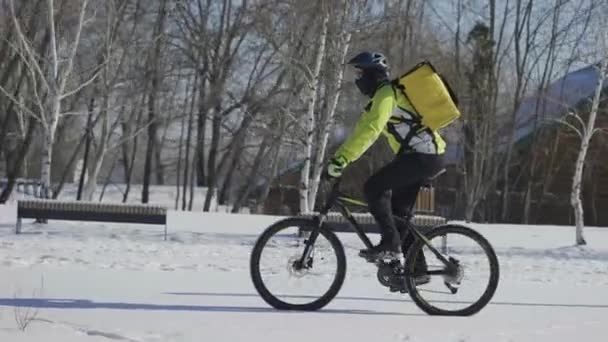 Entrega expressa de comida de bicicleta. Homem com mochila térmica amarela anda de bicicleta ao longo da rua coberta de neve no dia gelado. Ciclista em roupas quentes trabalha ao ar livre, servindo os clientes. Rastreamento. Movimento lento — Vídeo de Stock