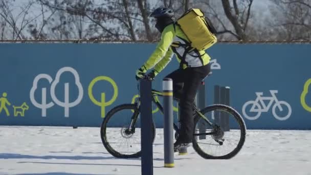 Entrega rápida de comida en bicicleta. El hombre con mochila termo amarilla monta en bicicleta a lo largo de la calle cubierta de nieve en un día helado. Ciclista en ropa de abrigo trabaja al aire libre, sirviendo a los clientes. Seguimiento. Movimiento lento — Vídeos de Stock