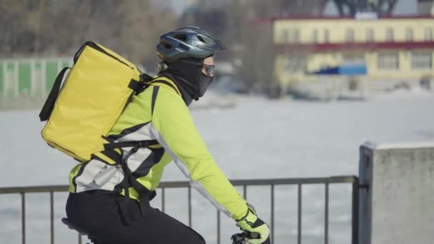 Homme avec un grand sac en casque promenades à vélo le long de la rue enneigée et livre de la nourriture ou des colis. Livraison rapide de courrier en ville. Conditions de travail extrêmes sur la route glissante d'hiver. Au ralenti. Gros plan. — Video