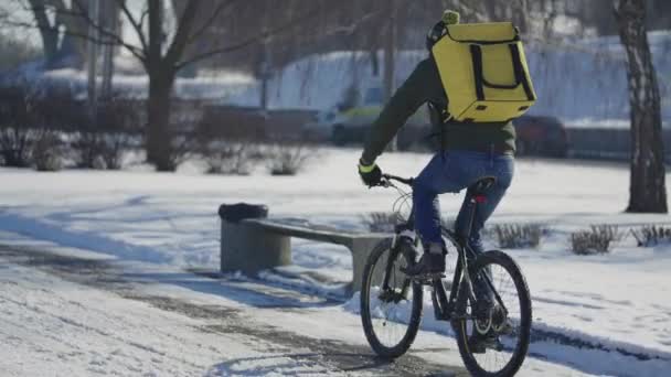 Vue arrière de l'homme avec un grand sac thermique jaune sur son dos fait du vélo le long de la rue enneigée et livre de la nourriture ou des colis. Livraison rapide par courrier à vélo. Paysage urbain enneigé. Mouvement lent. — Video