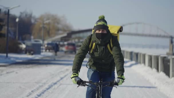 Portrait des Radfahrers mit Rucksack und Schutzmaske fährt Fahrrad auf schneebedeckter Straße mit Essensausgabe. Mann arbeitet als Kurier und liefert während des Virus-Ausbruchs Bestellungen an Kunden aus. Zeitlupe. — Stockvideo