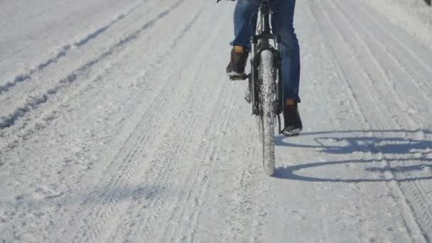 Hombre montar en bicicleta en un día nevado, condiciones de carretera resbaladizas. Cierre de pies de hombre pedaleando. Ruedas de bicicleta en la nieve. Servicio de mensajería de comida en bicicleta a lo largo de una calle cubierta de nieve. Movimiento lento. — Vídeo de stock