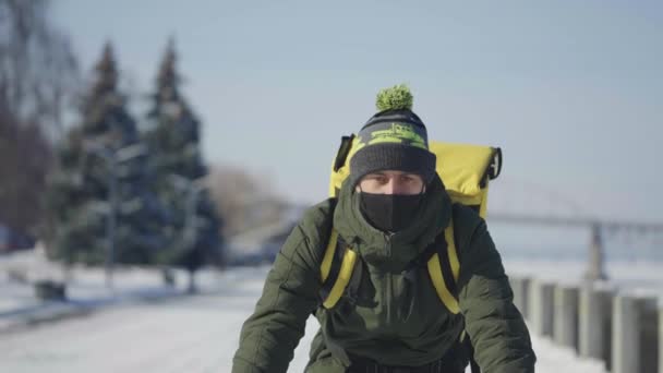 Cyklist med ryggsäck och skyddsmask cyklar på snötäckt gata med matleverans. Människan arbetar som kurir och levererar order till kunder vid virusutbrott. Sakta i backarna. Närbild. — Stockvideo