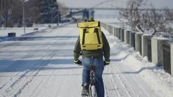 Visão traseira do homem com grande saco térmico amarelo em suas costas passeios de bicicleta ao longo da rua nevada e oferece comida ou parcelas. Entrega rápida de correio de bicicleta. Paisagem urbana nevada. Movimento lento. — Vídeo de Stock