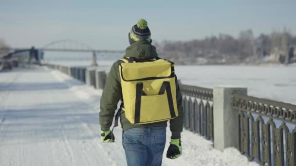 Widok z tyłu kuriera z dużą żółtą torbą termowizyjną spacerującą po pokrytej śniegiem ulicy miasta. Szybka dostawa jedzenia po mieście. Człowiek pracuje jako kurier i dostarcza zamówienia klientom. Zwolniony ruch. — Wideo stockowe