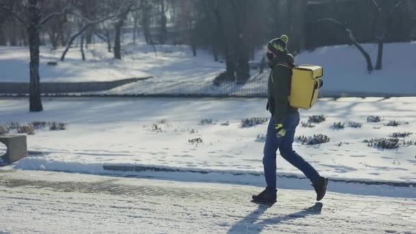 Oldalnézet futár orvosi maszk termo táska sétál a havas város utcájában. Gyors kaja szállítás a városban. Az ember futárként dolgozik, rendet szállít az ügyfeleknek a vírus kitörése alatt. Lassú mozgás.. — Stock videók