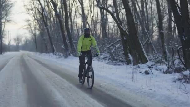 Велоспорт зимой. Велосипедист едет по скользкой асфальтовой дороге. Человек в шлеме и теплой спортивной одежде едет в зимний снежный лес в морозный день. Экстремальная прогулка по заснеженному лесу. Медленное движение. — стоковое видео