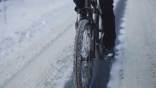 Fietsen in de winter door een besneeuwd bos. De fietser rijdt over een gladde asfaltweg. Mannelijke voeten trappen. Een close-up van een bewegende fiets. Extreem winterfietsen. Langzame beweging. — Stockvideo