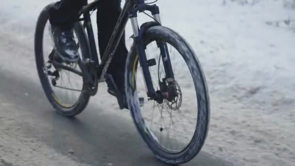 Cycling in the winter through a snowy forest. The cyclist rides on a slippery asphalt road. Male feet are pedaling. A close up of a moving bike. Extreme winter biking. Slow motion. — Stock Video