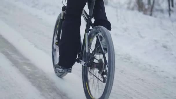Велоспорт зимой по заснеженному лесу. Велосипедист едет по скользкой асфальтовой дороге. Мужские ноги крутят педали. Крупный план движущегося велосипеда. Экстремальный зимний велосипед. Вид сзади. Медленное движение. — стоковое видео