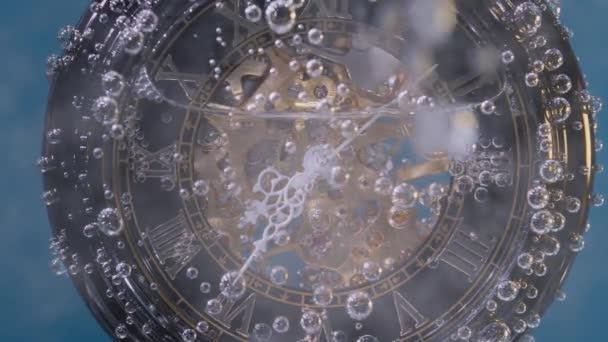 Reloj antiguo en una cadena cubierta de burbujas de aire en burbujeante agua transparente sobre un fondo azul. Primer plano de la esfera de un reloj girando desde la corriente de agua. Movimiento lento. — Vídeos de Stock