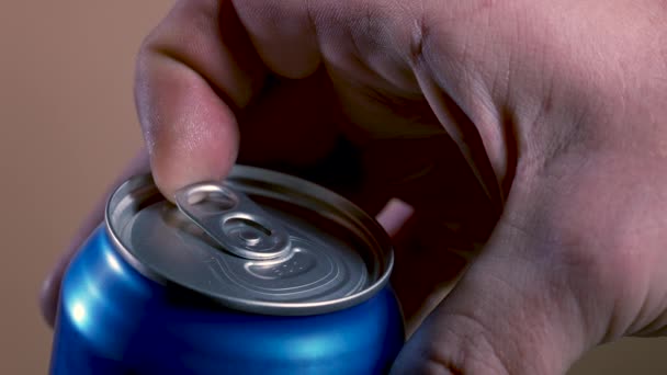 Ouverture d'une canette bleue secouée de bière, à partir de laquelle une boisson mousseuse est versée. Un plan détaillé d'une main d'homme ouvrant une canette de bière agitée sur un fond brun. Au ralenti. Gros plan. — Video