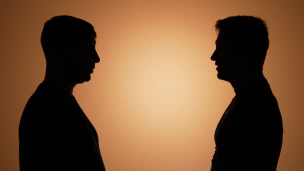 Os dois homens conversam, conversam, batem copos de garrafas de cerveja e bebem cerveja. Silhuetas de dois homens no estúdio em um fundo gradiente marrom. Conhecer amigos que gostam de bebidas. Movimento lento. Fechar. — Vídeo de Stock