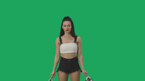 Сексуальна брюнетка в шортах махає чорно-білими картатими гоночними прапорами, щоб сигналізувати про початок змагань. Молода жінка позує на фоні зеленого екрану крупним планом. Повільний рух готовий, 4K на 59.94fps . — стокове відео