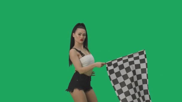 Morena sexy em shorts acenando preto e branco quadriculado bandeira de corrida para sinalizar o início da competição. Jovem posando contra fundo de tela verde fechar. Movimento lento pronto, 4K a 59.94fps. — Vídeo de Stock