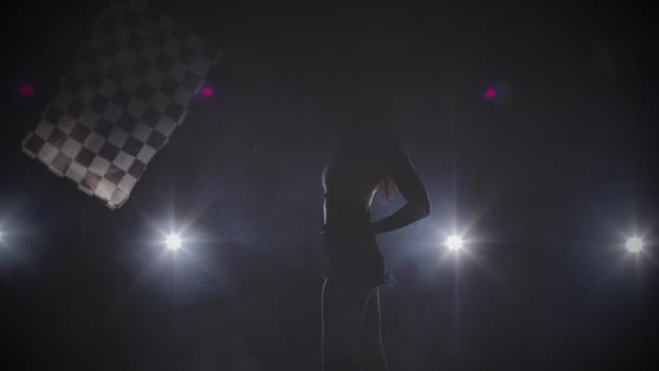 Widok z boku sylwetki młoda kobieta macha zaokrągloną flagą wyścigu, aby zasygnalizować początek imprezy wyścigowej. Brunetka pozująca w ciemnym studio z podświetleniem. Zamknij drzwi. Zwolnij, 4K przy 59,94fps.. — Wideo stockowe