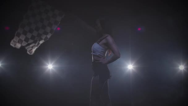 Zijaanzicht van silhouet jonge vrouw zwaaien geruite race vlag om het begin van race-evenement aan te geven. Brunette poseert in donkere rokerige studio met achtergrondverlichting. Sluit maar af. Slow motion klaar, 4K bij 59,94fps. — Stockvideo