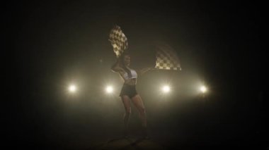 Güzel genç bir kadın yarış yarışmasında yarışırken kareli bayrak sallıyor. Esmer silueti, arka ışıklandırması olan dumanlı bir stüdyoda uzun süre poz veriyor. Yavaş çekim hazır, 4K 59.94fps 'de.
