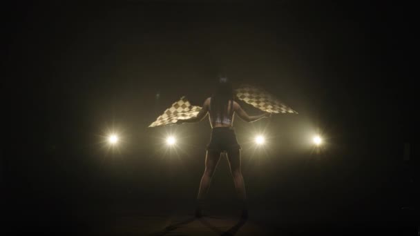 Young woman waving checkered flags while performing in racing competition. Back view silhouette of brunette poses full length in dark smoky studio with backlight. Slow motion ready, 4K at 59.94fps. — 图库视频影像