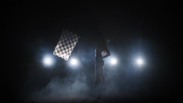 Zijaanzicht van silhouet jonge vrouw zwaaien geruite race vlag om het begin van een race-evenement aan te geven. Brunette poseert full length in donkere rokerige studio met achtergrondverlichting. Slow motion klaar, 4K bij 59,94fps — Stockvideo