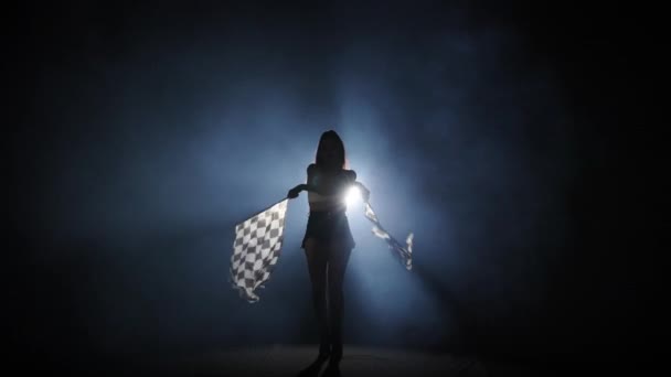 Silueta de mujer joven ondeando una bandera de carrera a cuadros para señalar el inicio de un evento de carreras. Morena posando de cuerpo entero en un oscuro estudio ahumado con contraluz. Cámara lenta lista, 4K a 59.94fps. — Vídeo de stock