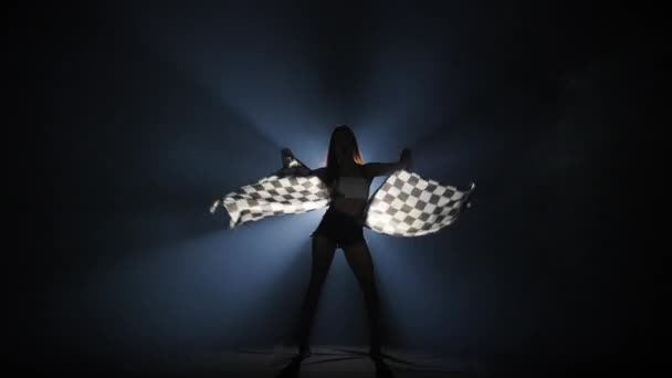 Silueta de mujer joven ondeando una bandera de carrera a cuadros para señalar el inicio de un evento de carreras. Morena posando de cuerpo entero en un oscuro estudio ahumado con contraluz. Cámara lenta lista, 4K a 59.94fps. — Vídeo de stock