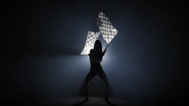 Vista trasera de la silueta joven mujer ondeando bandera de carrera a cuadros para señalar el inicio del evento de carreras. Morena posando de cuerpo entero en oscuro estudio ahumado con contraluz. Cámara lenta lista, 4K a 59.94fps. — Vídeo de stock