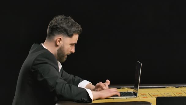 Side view of bearded man sitting at roulette table and playing in an online casino on laptop. Gentleman wins and celebrates victory. Money rain. Close up. Slow motion ready, 4K at 59.94fps. — Stock Video