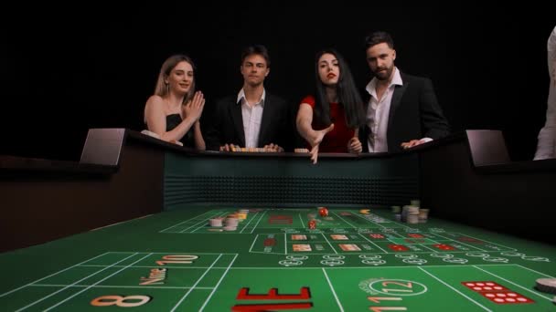 男性と女性のグループが緑の拍手テーブルに立っています。赤いドレスの女性はサイコロを転がして負けます。会社はとても動揺している。ゲームテーブル内からの眺め。スローモーション. — ストック動画