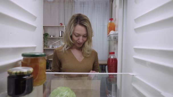 Mujer joven abre la puerta del refrigerador, sosteniendo bolsa de papel de comestibles y poniendo comida en la nevera. Vista desde el interior de la nevera. De cerca. Cámara lenta lista 59.94fps. — Vídeo de stock