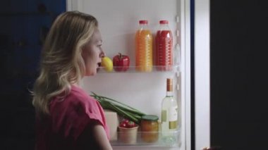 Genç ve güzel bir kadın geceleri buzdolabının kapısını açar, etrafa dikkatlice bakar ve çöreği ısırmaktan mutlu olur. Yan görüş. Kapatın. Yavaş çekim hazır 59.94fps.