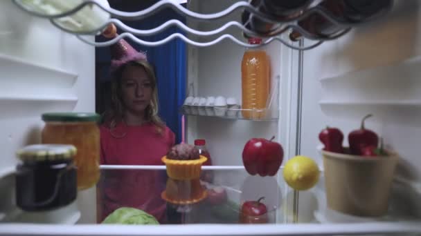 Eine einsame, aufgebrachte Frau mit festlicher Mütze holt nachts eine Torte mit Kerze aus dem Kühlschrank. Blick aus dem Inneren des Küchenkühlschranks. Aus nächster Nähe. Zeitlupe bereit 59.94fps. — Stockvideo