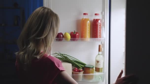 Молода жінка відкриває двері холодильника вночі, дивиться всередину і виймає капусту та огірки для салату. Здоровий спосіб життя, дієта. Вид збоку. Близько. Повільний рух готовий 59.94fps . — стокове відео