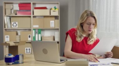 Depo çalışanı bir ofis odasında kutuların arka planına karşı çalışıyor. Dizüstü bilgisayarda yazan, tablet kullanan, paket teslimatları hakkında bilgi toplayan bir kadın. Kapatın. Yavaş çekim hazır 59.94fps.