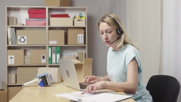 Trabajadora de correos con auriculares trabajando en la computadora portátil en la oficina de correos y tecleando en el teclado. Mujer joven sentada a la mesa sobre fondo de estantes con cajas. De cerca. Cámara lenta lista 59.94fps. — Vídeo de stock