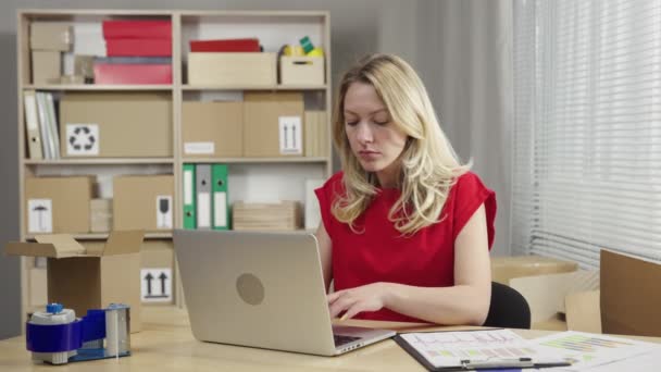 Pakhuismedewerker werkt in een kantoorruimte tegen de achtergrond van dozen. Vrouw typt op laptop, dan verheugt zich een goede deal op de levering pakketten. Sluit maar af. Slow motion klaar 59,94fps. — Stockvideo