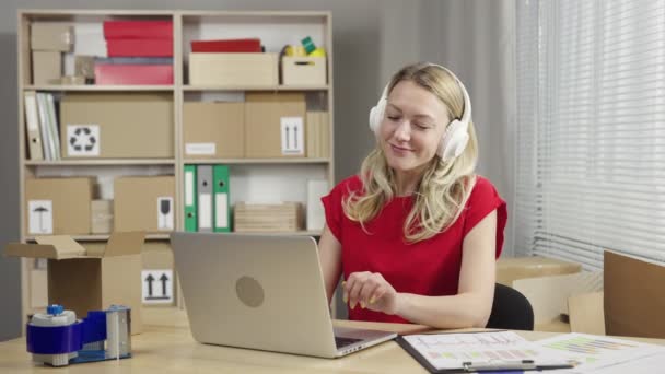 Női postás gépel laptopon, és élvezi a zenét nagy fehér fejhallgatóval. Egy nő ül az asztalnál a polcok mögött dobozokkal a postahivatalban. Közelről. Lassú mozgás kész 59.94fps — Stock videók