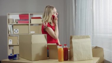 Depo çalışanı bir ofis odasında kutuların arka planına karşı çalışıyor. Kadın müşterilere teslimat için paketler hazırlıyor ve akıllı telefondan konuşuyor. Kapatın. Yavaş çekim hazır 59.94fps.