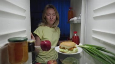 Genç kadın geceleri buzdolabının kapısını açıyor ve hamburger yerine elma alıyor. Sağlıklı yaşam tarzı, diyet. Çalışan buzdolabının içinden bak. Kapatın. Yavaş çekim hazır 59.94fps.