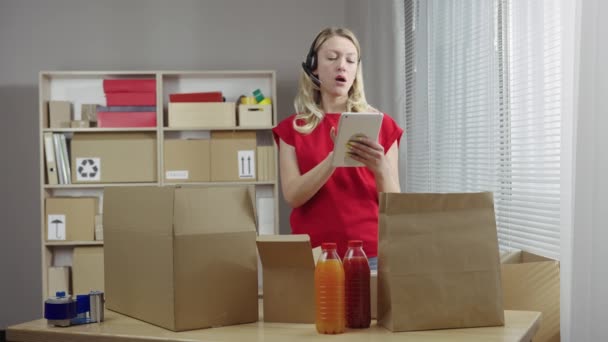 A raktár dolgozója egy irodai szobában dolgozik dobozok háttere ellenére. A nő egy fülhallgató és egy tabletta segítségével készíti el a vevőknek történő kiszállításra szánt élelmiszercsomagokat. Közelről. Lassú mozgás kész 59.94fps. — Stock videók