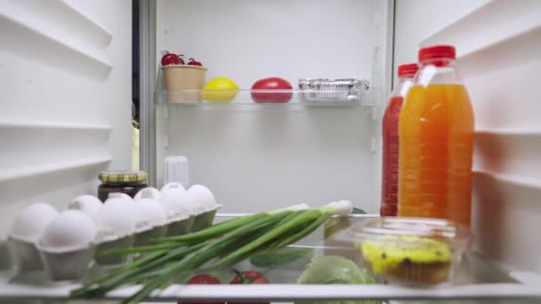 Mulher jovem e sonolenta abre a porta da geladeira à noite, olha para dentro e escolhe suco de laranja. Vista de dentro da geladeira da cozinha. Fecha. Movimento lento pronto 59.94fps. — Vídeo de Stock