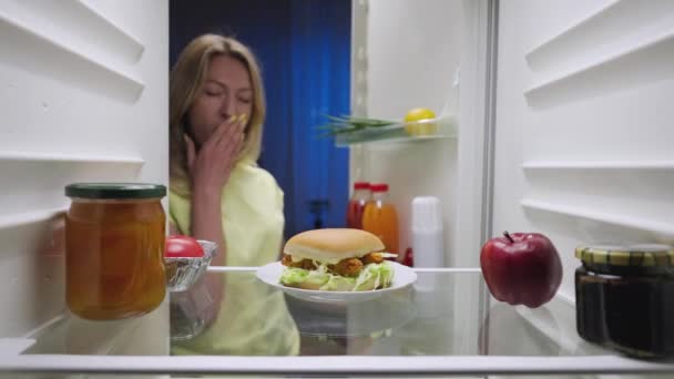 Uykulu genç kadın geceleri buzdolabının kapısını açar, etrafa dikkatlice bakar ve mutlu bir şekilde hamburgeri alır. Mutfak dolabının içinden bak. Kapatın. Yavaş çekim hazır 59.94fps. — Stok video