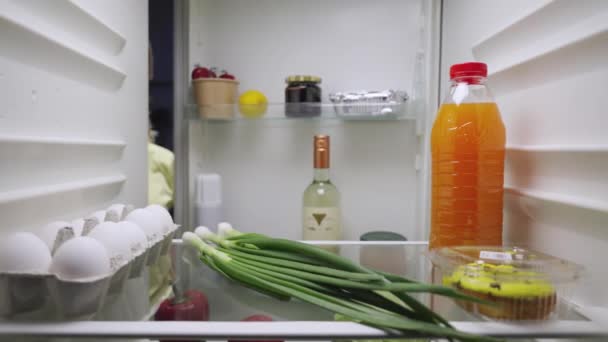 Νυσταγμένη νεαρή γυναίκα ανοίγει την πόρτα του ψυγείου το βράδυ, κοιτάζει μέσα και διαλέγει ένα μπουκάλι λευκό κρασί. Θέα μέσα από το ψυγείο της κουζίνας. Κλείσε. Αργή κίνηση έτοιμη 59.94fps. — Αρχείο Βίντεο
