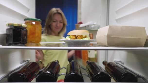 Mujer joven abre la puerta del refrigerador por la noche, mira dentro y feliz saca dos botellas de cerveza. Vista desde el interior de la nevera de la cocina. De cerca. Cámara lenta lista 59.94fps. — Vídeo de stock