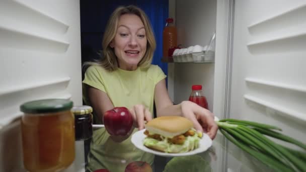 Jeune femme ouvre la porte du réfrigérateur la nuit et heureux prend un hamburger au lieu d'une pomme. Vue de l'intérieur du réfrigérateur de travail. Ferme là. Ralenti prêt 59.94fps. — Video