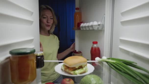 Eine junge Frau öffnet nachts die Kühlschranktür und nimmt glücklich einen Burger. Blick von innen auf den funktionierenden Kühlschrank. Aus nächster Nähe. Zeitlupe bereit 59.94fps. — Stockvideo