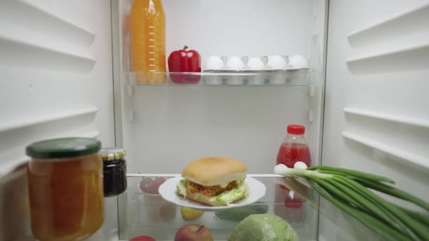 Jovem abre a porta da geladeira à noite e feliz leva um hambúrguer. Vista de dentro do frigorífico de trabalho. Fecha. Movimento lento pronto 59.94fps. — Vídeo de Stock